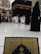 The image depicts a busy scene near the Kaaba in Mecca, with numerous people dressed in traditional pilgrimage attire, such as white robes for men and black coverings for women. In the foreground, a book with ornate gold and black patterns is partially visible, possibly a Quran. Surrounding the Kaaba are large buildings with modern architectural design.