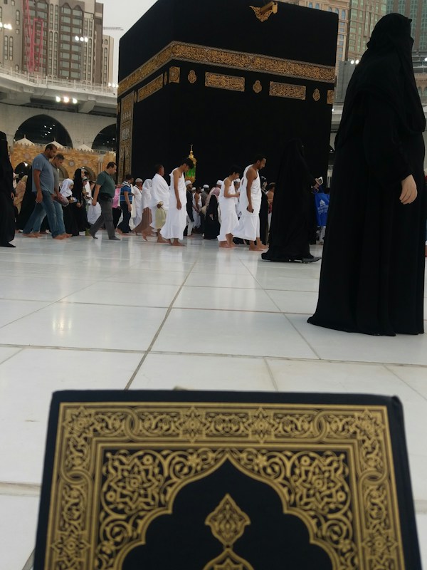 The image depicts a busy scene near the Kaaba in Mecca, with numerous people dressed in traditional pilgrimage attire, such as white robes for men and black coverings for women. In the foreground, a book with ornate gold and black patterns is partially visible, possibly a Quran. Surrounding the Kaaba are large buildings with modern architectural design.