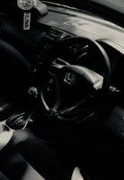 The interior of a car featuring a steering wheel with the Honda logo, gear shift, and part of a dashboard. A car air freshener hangs from the rearview mirror.