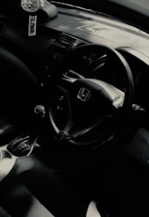 The interior of a car featuring a steering wheel with the Honda logo, gear shift, and part of a dashboard. A car air freshener hangs from the rearview mirror.