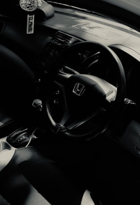 The interior of a car featuring a steering wheel with the Honda logo, gear shift, and part of a dashboard. A car air freshener hangs from the rearview mirror.