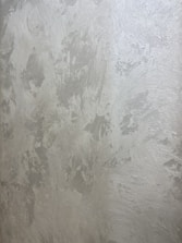 A textured wall with a plaster-like appearance, featuring a mix of light gray and subtle warm tones. The surface displays a variety of brushstroke patterns, creating a rustic and uneven visual effect.