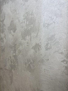 A textured wall with a plaster-like appearance, featuring a mix of light gray and subtle warm tones. The surface displays a variety of brushstroke patterns, creating a rustic and uneven visual effect.