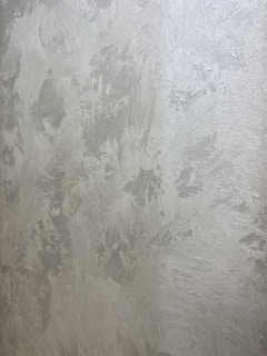 A textured wall with a plaster-like appearance, featuring a mix of light gray and subtle warm tones. The surface displays a variety of brushstroke patterns, creating a rustic and uneven visual effect.