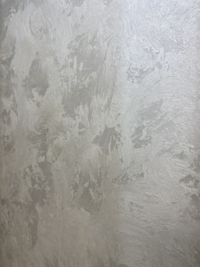 A textured wall with a plaster-like appearance, featuring a mix of light gray and subtle warm tones. The surface displays a variety of brushstroke patterns, creating a rustic and uneven visual effect.