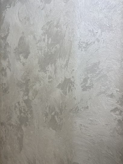 A textured wall with a plaster-like appearance, featuring a mix of light gray and subtle warm tones. The surface displays a variety of brushstroke patterns, creating a rustic and uneven visual effect.