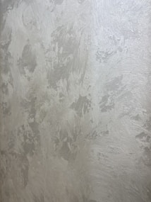 A textured wall with a plaster-like appearance, featuring a mix of light gray and subtle warm tones. The surface displays a variety of brushstroke patterns, creating a rustic and uneven visual effect.