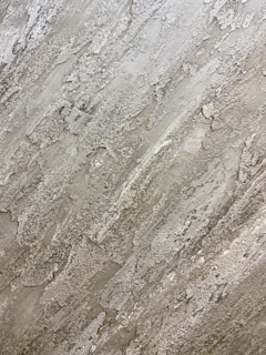 A textured surface with a rough, naturalistic pattern that resembles stone or concrete. The surface features various shades of gray with patches of lighter and darker areas, creating a rugged, layered effect.