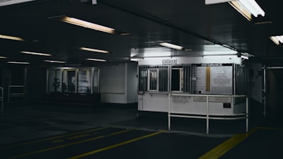 A dimly lit interior features a cashier booth with bars and signage. The space appears industrial, with fluorescent lighting casting harsh shadows. Information and notices are displayed on the wall behind the cashier booth.