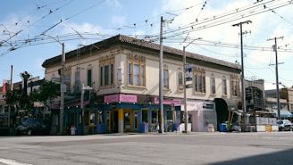 A two-story building with ornate architectural details, featuring a mix of businesses on the ground floor, including a taco bar and art bar. The street is lined with overhead wires and utility poles. There is a car parked on the side of the road, and a mix of trees and street signs are visible.