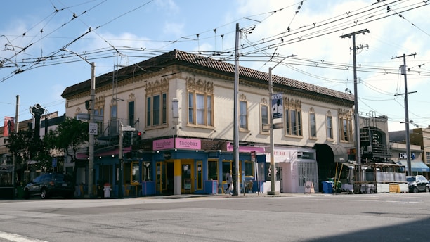 A two-story building with ornate architectural details, featuring a mix of businesses on the ground floor, including a taco bar and art bar. The street is lined with overhead wires and utility poles. There is a car parked on the side of the road, and a mix of trees and street signs are visible.