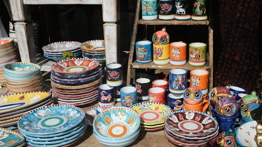 A display of colorful, hand-painted ceramic dishes and mugs on a wooden stand. The ceramics feature vibrant patterns and illustrations, including animal motifs and intricate designs. Several stacks of plates and bowls are arranged alongside mugs and owl-shaped figurines, showcasing a variety of artistic styles.