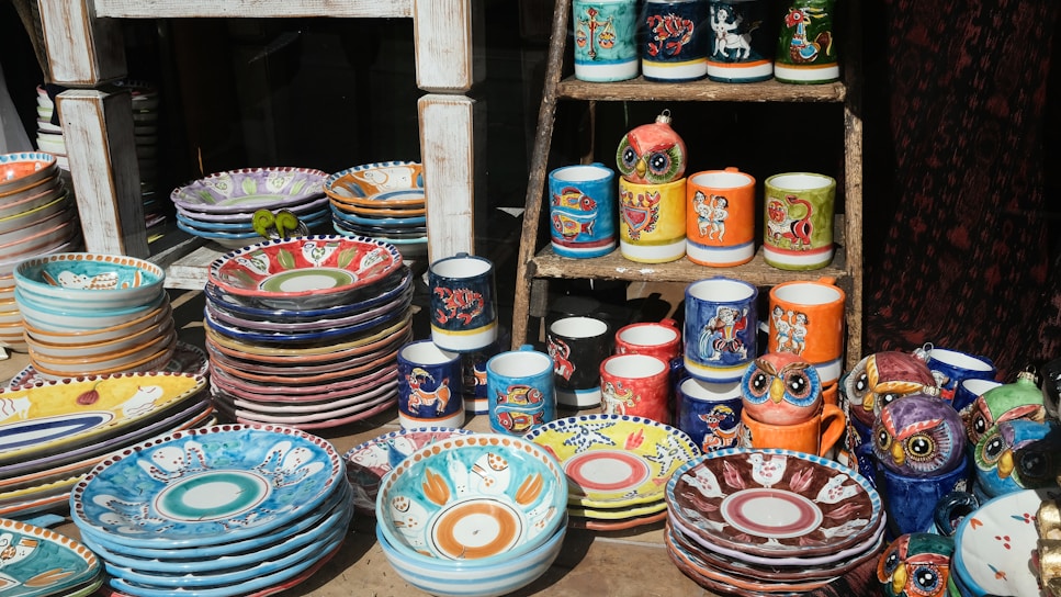A display of colorful, hand-painted ceramic dishes and mugs on a wooden stand. The ceramics feature vibrant patterns and illustrations, including animal motifs and intricate designs. Several stacks of plates and bowls are arranged alongside mugs and owl-shaped figurines, showcasing a variety of artistic styles.