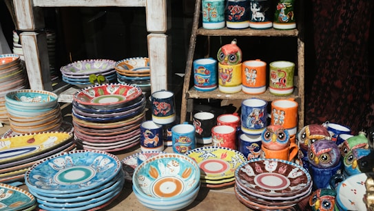 A display of colorful, hand-painted ceramic dishes and mugs on a wooden stand. The ceramics feature vibrant patterns and illustrations, including animal motifs and intricate designs. Several stacks of plates and bowls are arranged alongside mugs and owl-shaped figurines, showcasing a variety of artistic styles.