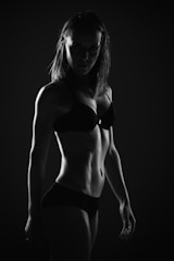A monochrome image of a person in underwear, illuminated from the back and side, highlighting the muscular contours of the body in a dramatic pose. The background is dark, adding to the contrast and emphasizing the silhouette.