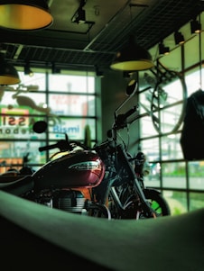 A motorcycle with a shiny red fuel tank is prominently displayed against a backdrop of large windows. Natural light filters through glass panes, casting an industrial ambiance with overhead lights and exposed ceiling structures. The setting appears to be a showroom or garage with a modern, urban feel.