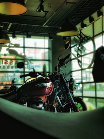 A motorcycle with a shiny red fuel tank is prominently displayed against a backdrop of large windows. Natural light filters through glass panes, casting an industrial ambiance with overhead lights and exposed ceiling structures. The setting appears to be a showroom or garage with a modern, urban feel.