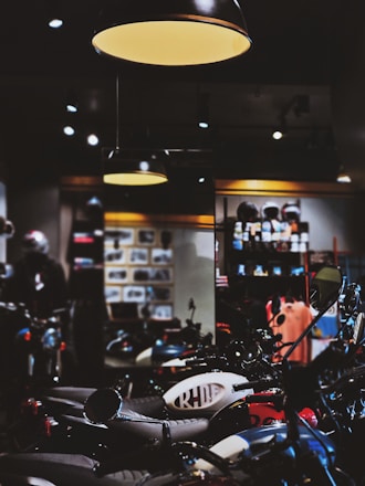 A dimly lit motorcycle showroom features several motorcycles parked in rows. Overhead lights cast a warm glow, highlighting the bikes' details. The background is filled with shelves displaying helmets and various motorcycle-related items.