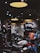 A dimly lit motorcycle showroom features several motorcycles parked in rows. Overhead lights cast a warm glow, highlighting the bikes' details. The background is filled with shelves displaying helmets and various motorcycle-related items.