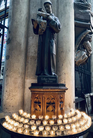 A statue depicting a saint holding a child is displayed on a pedestal with religious motifs. Surrounding the base are numerous lit candles arranged in a semicircle, casting a warm glow on the scene. The background features tall stone columns and stained glass windows, suggesting a church setting.