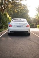 A luxury sedan is parked on a tree-lined street with its rear facing the viewer. The car is white with a sleek design, featuring modern taillights and dual exhaust pipes. The surroundings include lush green foliage and a well-maintained road.