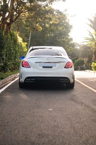 A luxury sedan is parked on a tree-lined street with its rear facing the viewer. The car is white with a sleek design, featuring modern taillights and dual exhaust pipes. The surroundings include lush green foliage and a well-maintained road.