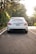 A luxury sedan is parked on a tree-lined street with its rear facing the viewer. The car is white with a sleek design, featuring modern taillights and dual exhaust pipes. The surroundings include lush green foliage and a well-maintained road.