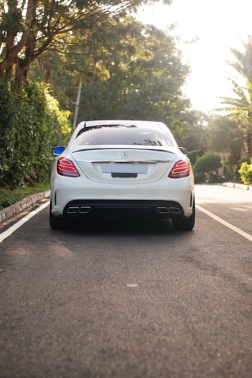 A luxury sedan is parked on a tree-lined street with its rear facing the viewer. The car is white with a sleek design, featuring modern taillights and dual exhaust pipes. The surroundings include lush green foliage and a well-maintained road.