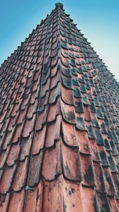A close-up view of a steeply angled roof covered with weathered red terracotta tiles, arranged in a patterned fashion. A wire or cable runs vertically along the tiles, secured with small metal fasteners. The sky is clear with a light blue gradient in the background.