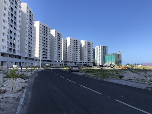 A series of tall, white residential apartment buildings lines the left side of a paved road. Construction is ongoing in the area with visible construction materials and a crane in the background. Sparse greenery is present on either side of the road leading up to the buildings.