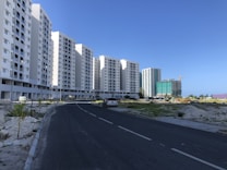 A series of tall, white residential apartment buildings lines the left side of a paved road. Construction is ongoing in the area with visible construction materials and a crane in the background. Sparse greenery is present on either side of the road leading up to the buildings.