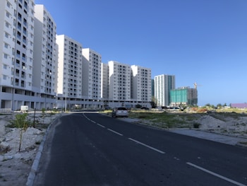 A series of tall, white residential apartment buildings lines the left side of a paved road. Construction is ongoing in the area with visible construction materials and a crane in the background. Sparse greenery is present on either side of the road leading up to the buildings.