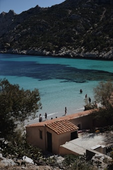 A picturesque coastal scene features turquoise waters gently lapping against a sandy beach. People are leisurely walking and standing in the shallow waters, taking in the serene environment. A rustic building with a terracotta roof sits nestled among lush greenery. Rugged hills and rocky cliffs rise in the background under a clear blue sky.