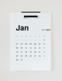 A minimalist wall calendar displaying the month of January 2023. The calendar features a simple design with bold black lettering for the days of the week and numbers representing the dates in a sans-serif font. The weekends are highlighted in a brownish-orange color. The calendar is hung on a white background, suggesting a clean and organized space.