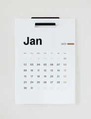 A minimalist wall calendar displaying the month of January 2023. The calendar features a simple design with bold black lettering for the days of the week and numbers representing the dates in a sans-serif font. The weekends are highlighted in a brownish-orange color. The calendar is hung on a white background, suggesting a clean and organized space.