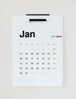 A minimalist wall calendar displaying the month of January 2023. The calendar features a simple design with bold black lettering for the days of the week and numbers representing the dates in a sans-serif font. The weekends are highlighted in a brownish-orange color. The calendar is hung on a white background, suggesting a clean and organized space.