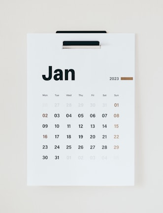 A minimalist wall calendar displaying the month of January 2023. The calendar features a simple design with bold black lettering for the days of the week and numbers representing the dates in a sans-serif font. The weekends are highlighted in a brownish-orange color. The calendar is hung on a white background, suggesting a clean and organized space.