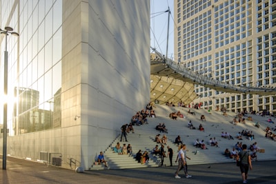 Large modern architectural structure with a wide set of steps where numerous people are sitting and socializing. The building has a distinctive, geometric design with extensive glass and metal elements. Sunlight casts reflections on the glass facade, and the scene is bustling with activity.