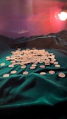 A collection of gold coins is spread out on a lush, dark green velvet cloth. The background is softly illuminated with a warm light, casting subtle reflections and shadows over the scene.
