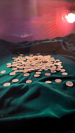A collection of gold coins is spread out on a lush, dark green velvet cloth. The background is softly illuminated with a warm light, casting subtle reflections and shadows over the scene.