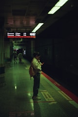 A person stands on a dimly lit subway platform looking at their phone. Nearby, an overhead electronic display shows information about train schedules. The platform has directional markings and is surrounded by a dark tunnel.