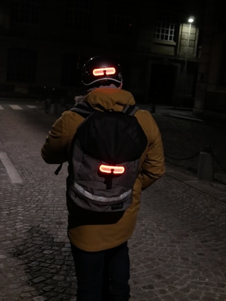 A person wearing a yellow jacket and a backpack is walking on a dimly lit cobblestone street at night. The person is wearing a helmet with a red light on the back and the backpack also has a red light attached to it, enhancing visibility in the darkness. The surroundings are dim with a few lit windows and a streetlamp in the background.