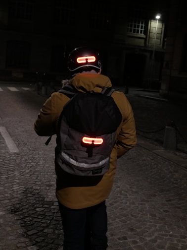 A person wearing a yellow jacket and a backpack is walking on a dimly lit cobblestone street at night. The person is wearing a helmet with a red light on the back and the backpack also has a red light attached to it, enhancing visibility in the darkness. The surroundings are dim with a few lit windows and a streetlamp in the background.