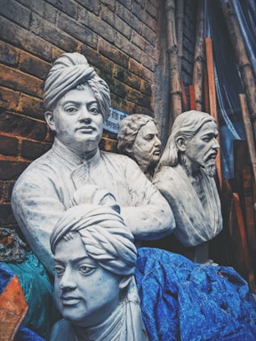 Four realistic white marble-like busts are displayed against a brick wall backdrop. The sculptures appear intricately carved, capturing detailed facial expressions and features. They are partially covered with blue fabric, and there are bamboo sticks leaning against the wall.