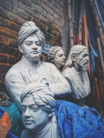 Four realistic white marble-like busts are displayed against a brick wall backdrop. The sculptures appear intricately carved, capturing detailed facial expressions and features. They are partially covered with blue fabric, and there are bamboo sticks leaning against the wall.