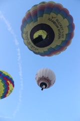 Multiple hot air balloons of varying colors float against a clear blue sky. One balloon features a rainbow pattern, while another has geometric designs. A visible flame heats the air in the largest balloon.