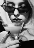 A black and white photograph featuring a person with platinum blonde hair and dark sunglasses holding a cocktail in a martini glass. The individual has bold lipstick and neatly manicured nails. The image exudes an air of sophistication and mystery.
