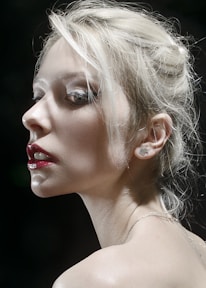 A dramatic portrait of a person with platinum blonde hair in an updo, accentuated by deep red lipstick and subtle makeup. The lighting highlights the contours of the face, casting soft shadows and creating a mysterious and sophisticated atmosphere.