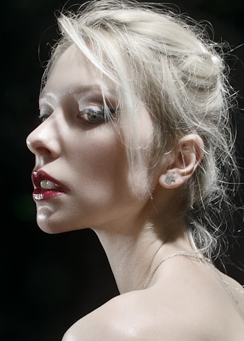 A dramatic portrait of a person with platinum blonde hair in an updo, accentuated by deep red lipstick and subtle makeup. The lighting highlights the contours of the face, casting soft shadows and creating a mysterious and sophisticated atmosphere.