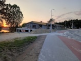 A construction site with a building being erected near a paved road, surrounded by temporary fencing. The scene is set during sunset, evident from the golden glow in the sky. Trees and shrubbery appear in the background, with a notable large tree on the left side. There are mounds of earth and building materials around the site, and the road curves away into the distance.